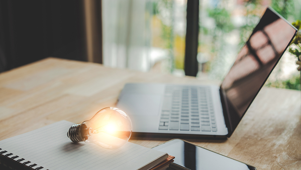 A lightbulb next to a laptop, smartphone, and writing pad
